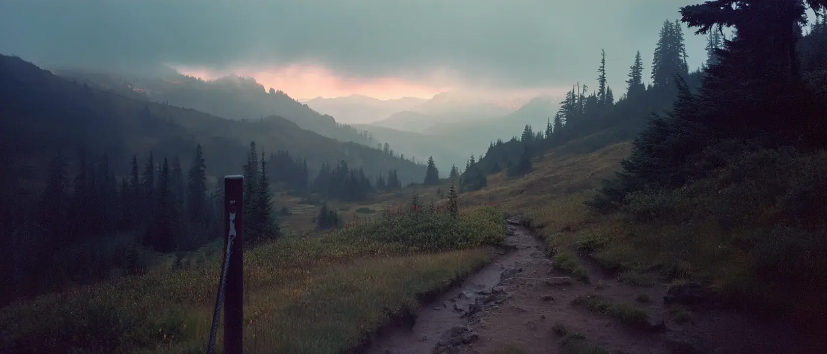 A dirt trail leading from a marker post into a misty mountain valley at first light